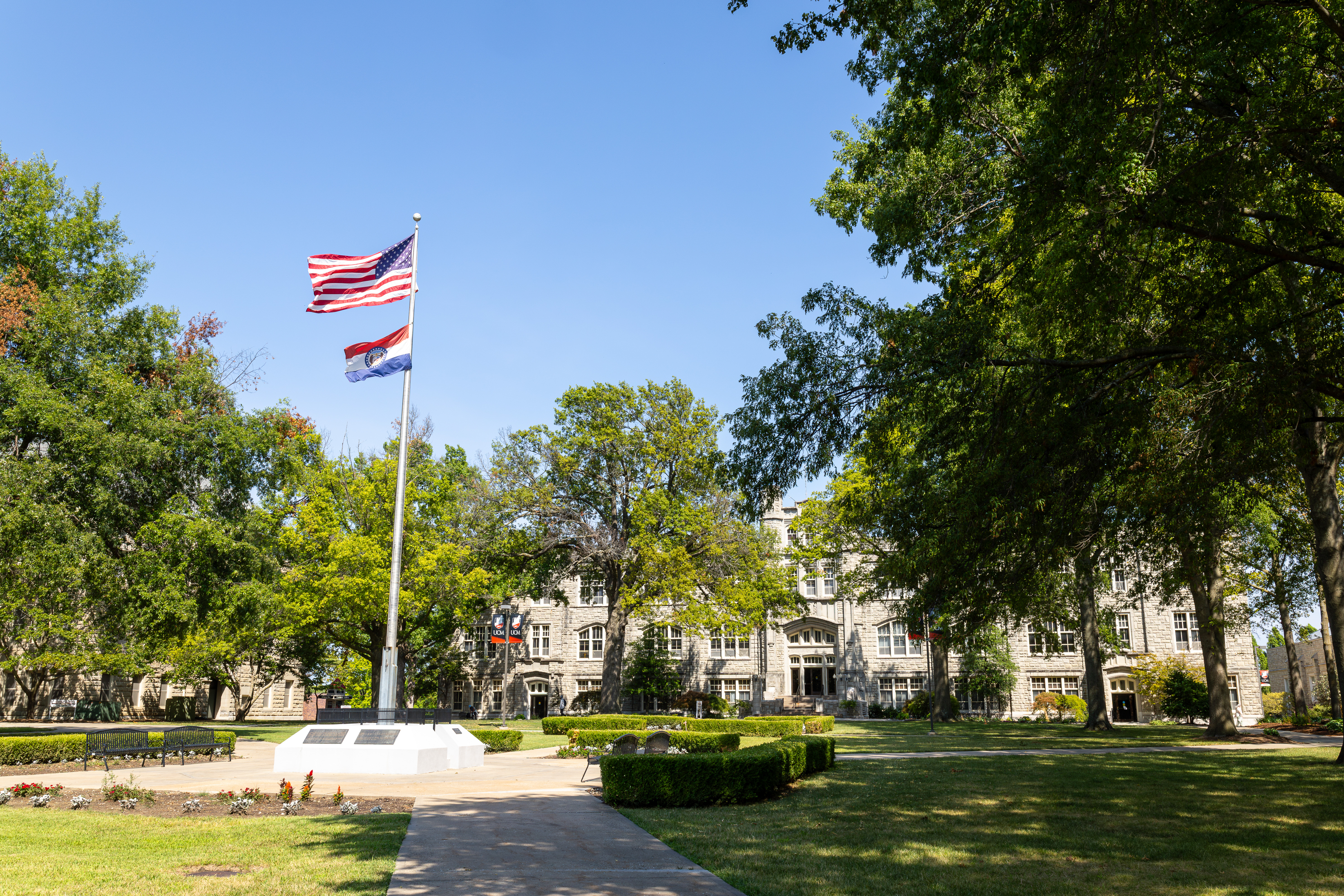 A tall flagpole in the middle of the UCM quad has an American flag and Missouri flag waving in the wind. Green trees and grass are seen, with a large stone building in the background