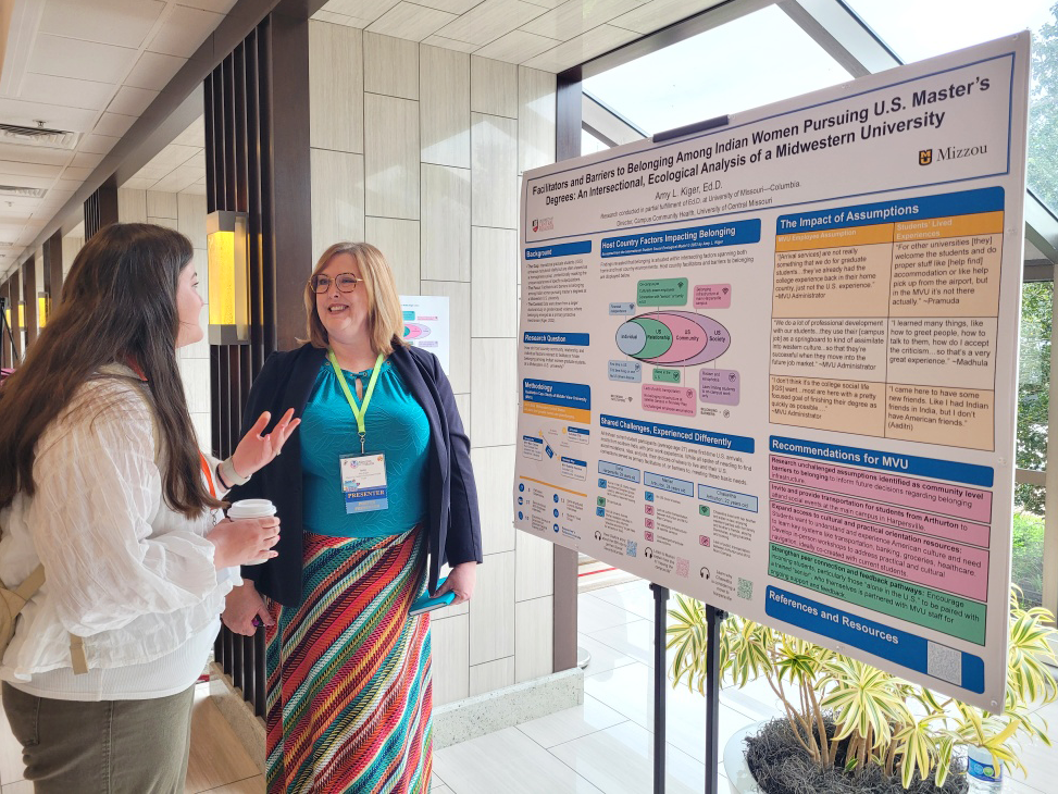 Two people talk to each other in a hallway while standing in front of a large poster, which is in front of a wall of windows. The poster has a lot of text and colorful charts about the research being presented.