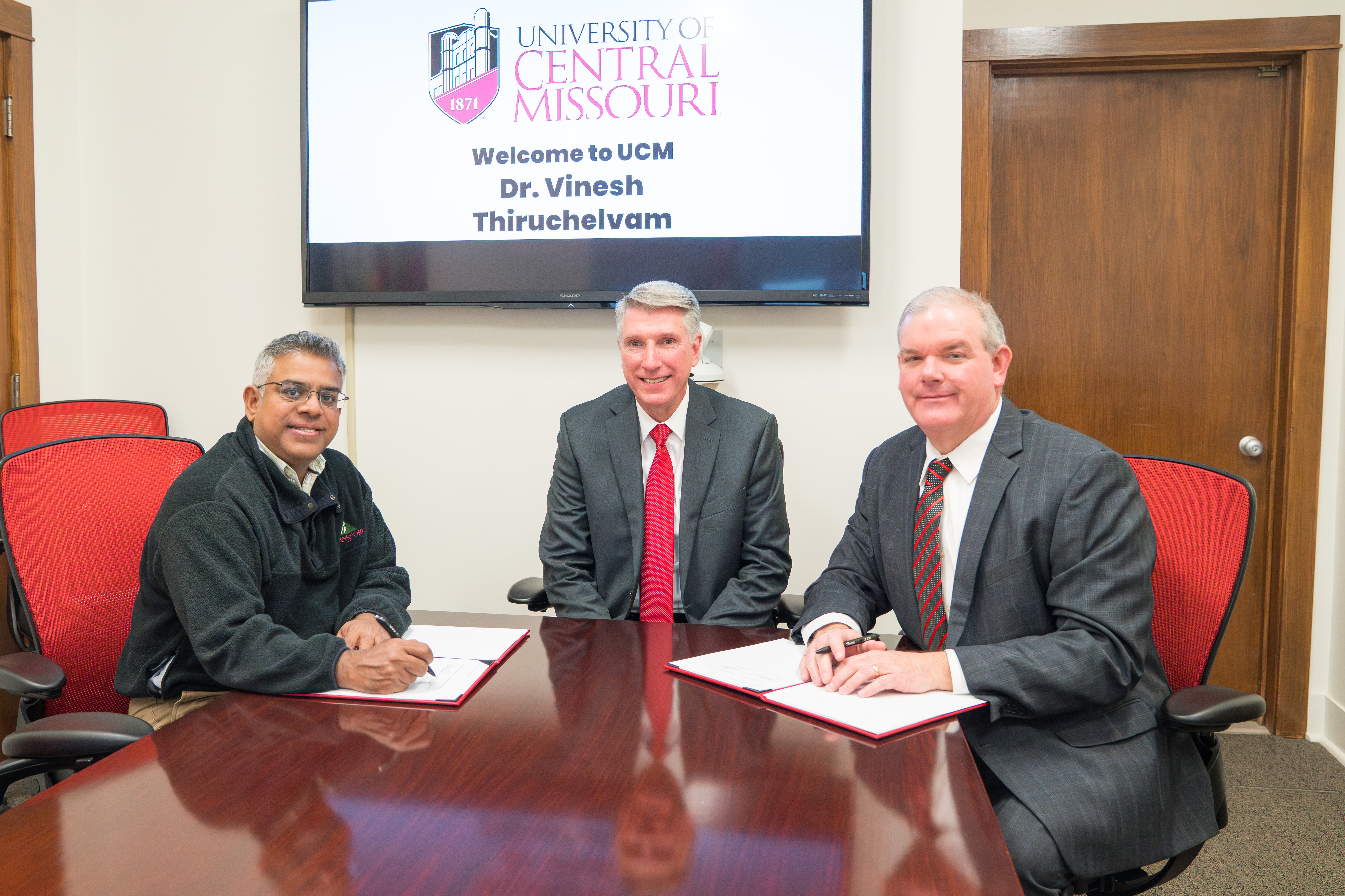Three people sit at a wood table in a conference room with a TV on the wall behind them. They are smiling and looking at the camera, and two have documents in front of them to sign.