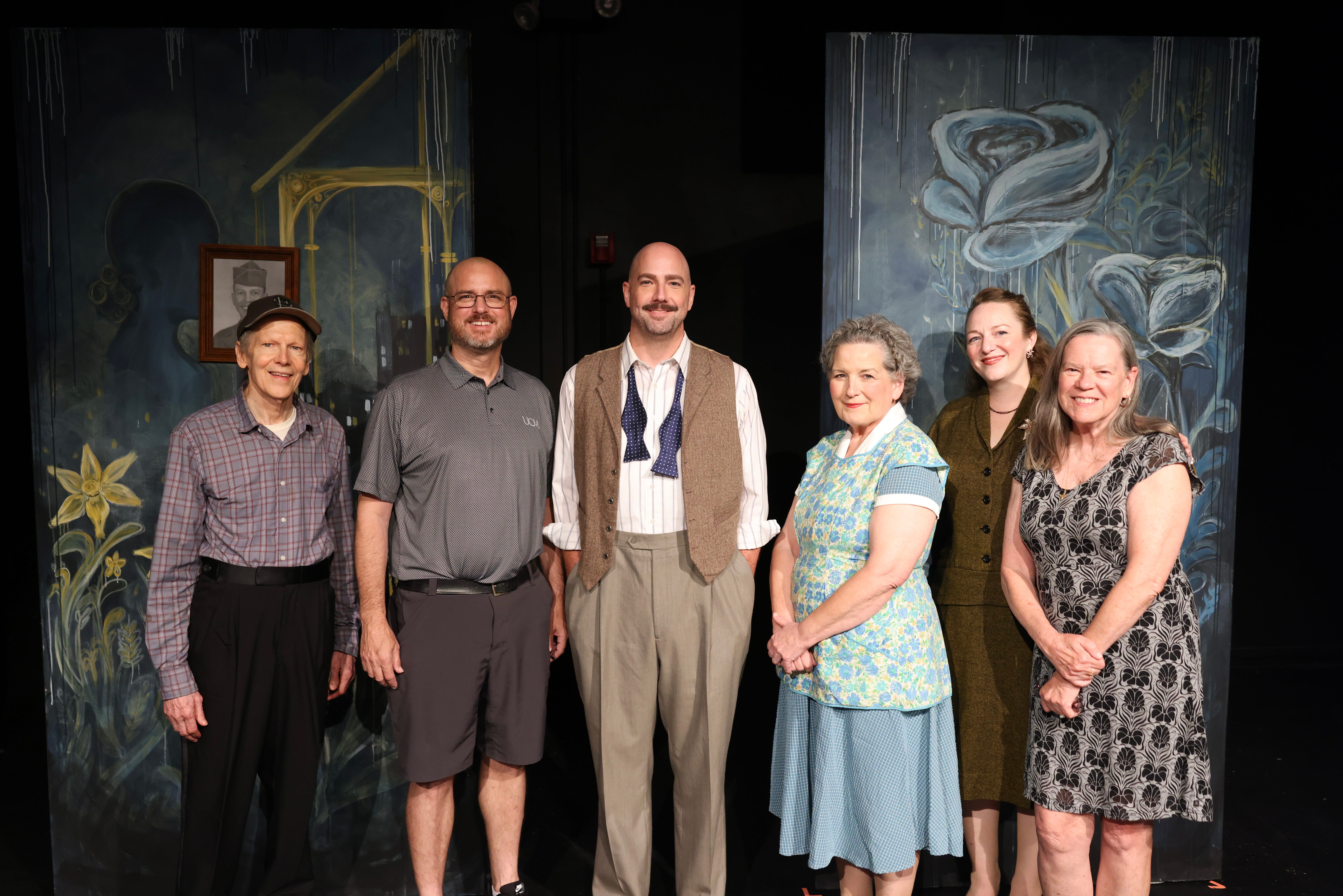 Six people stand next to each other on a theatre stage with a backdrop for the play behind them. Some of the people are actors who are in costume for the 1940s-era play, while Aaron Scully is wearing a gray UCM polo and black shorts and the director is wearing a black-and-gray patterened dress. They are all looking at the camera and smiling.