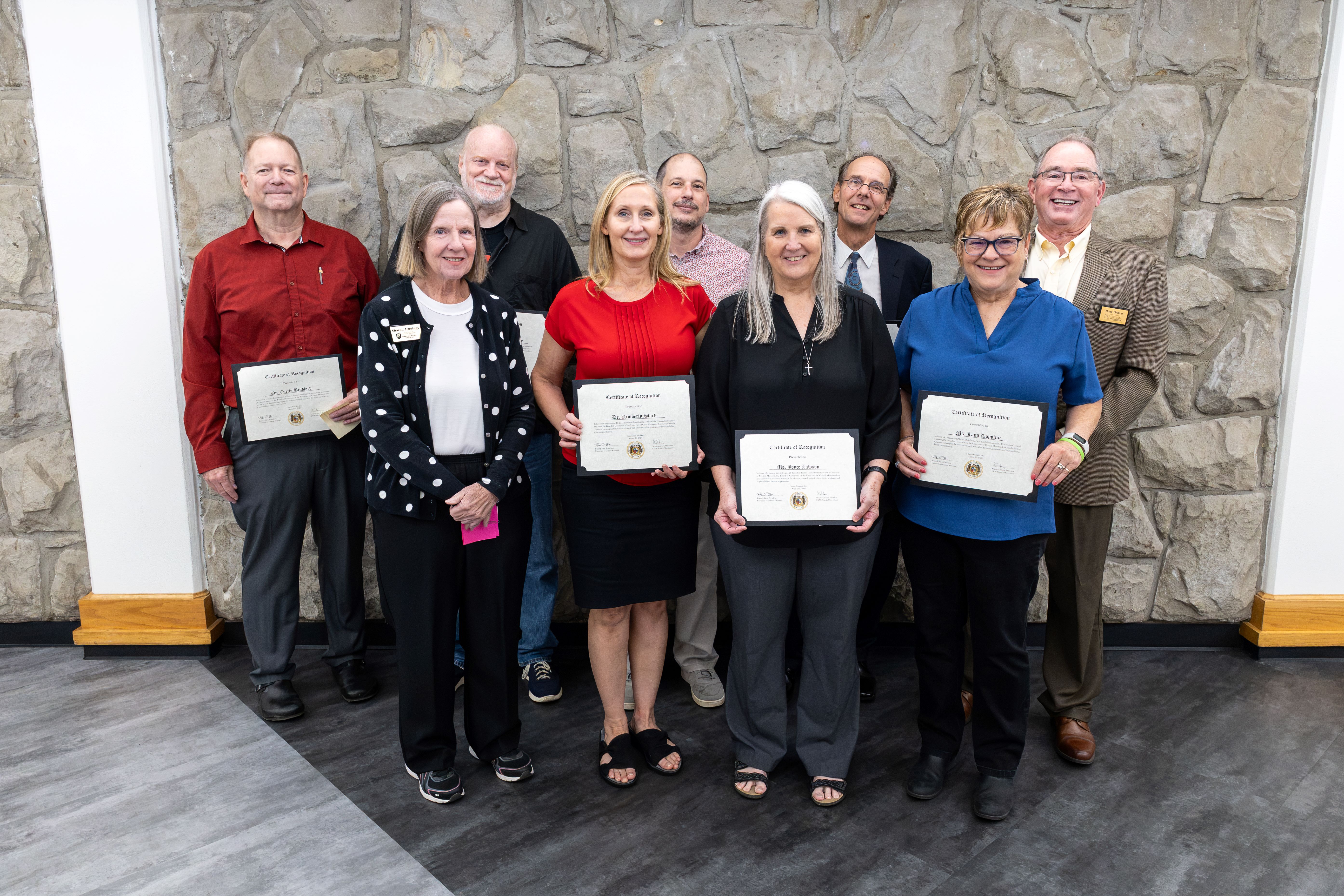 Four adults stand in the front row and five stand in the back row. They are smiling and looking at the camera while standing in a hallway in front of a gray stone wall. Several people are holding certificates.