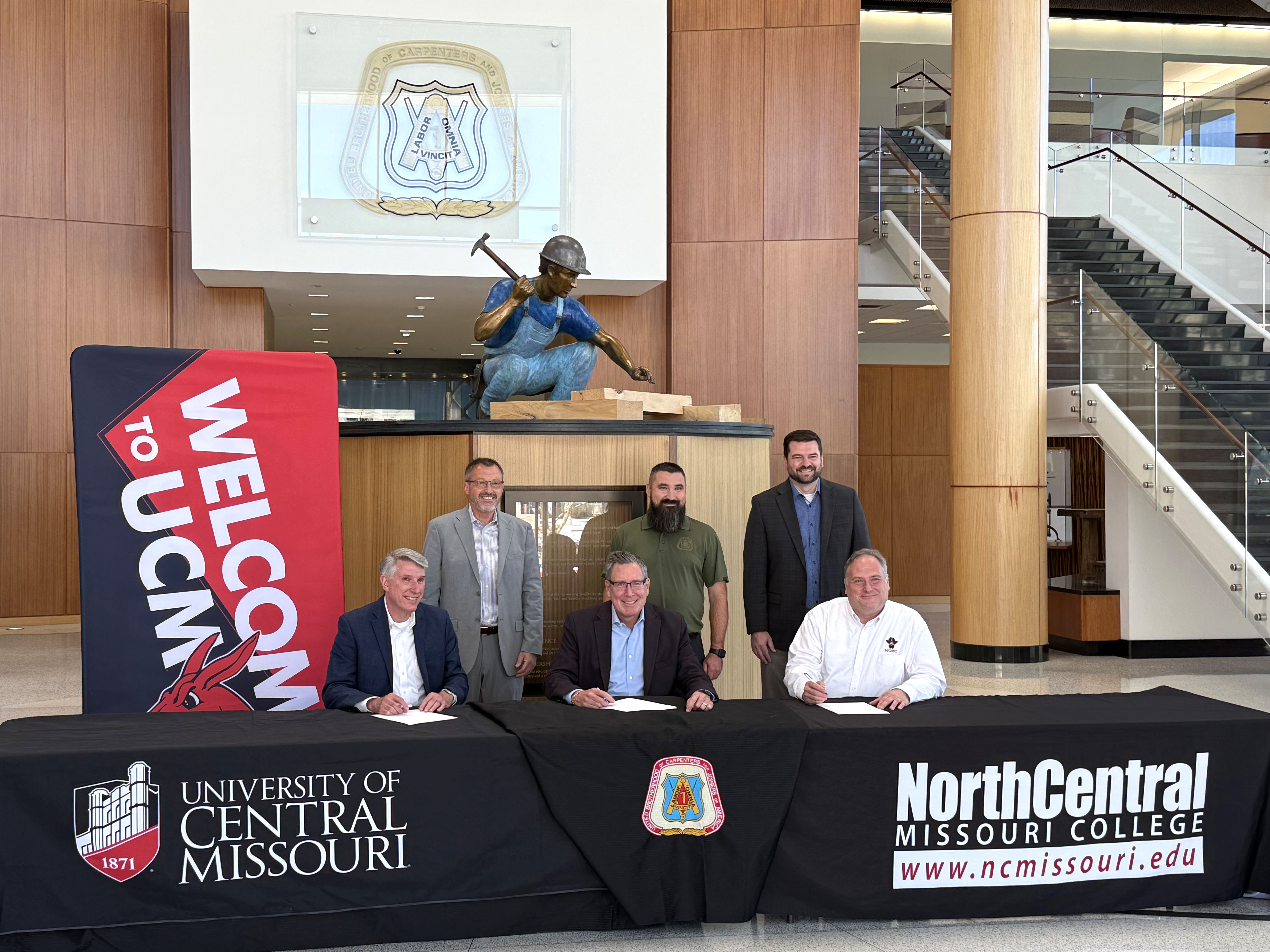 Three people are sitting and three people are standing behind a long table in the foyer of the Carpenters International Training Center. Everyone is smiling and looking at the photo. The three people sitting are preparing to sign paperwork. One black tablecloth says “University of Central Missouri,” one says “North Central Missouri College www.ncmissouri.edu” and the third has the United Brotherhood of Carpenters logo. The people are in front of a statue of a carpenter. 