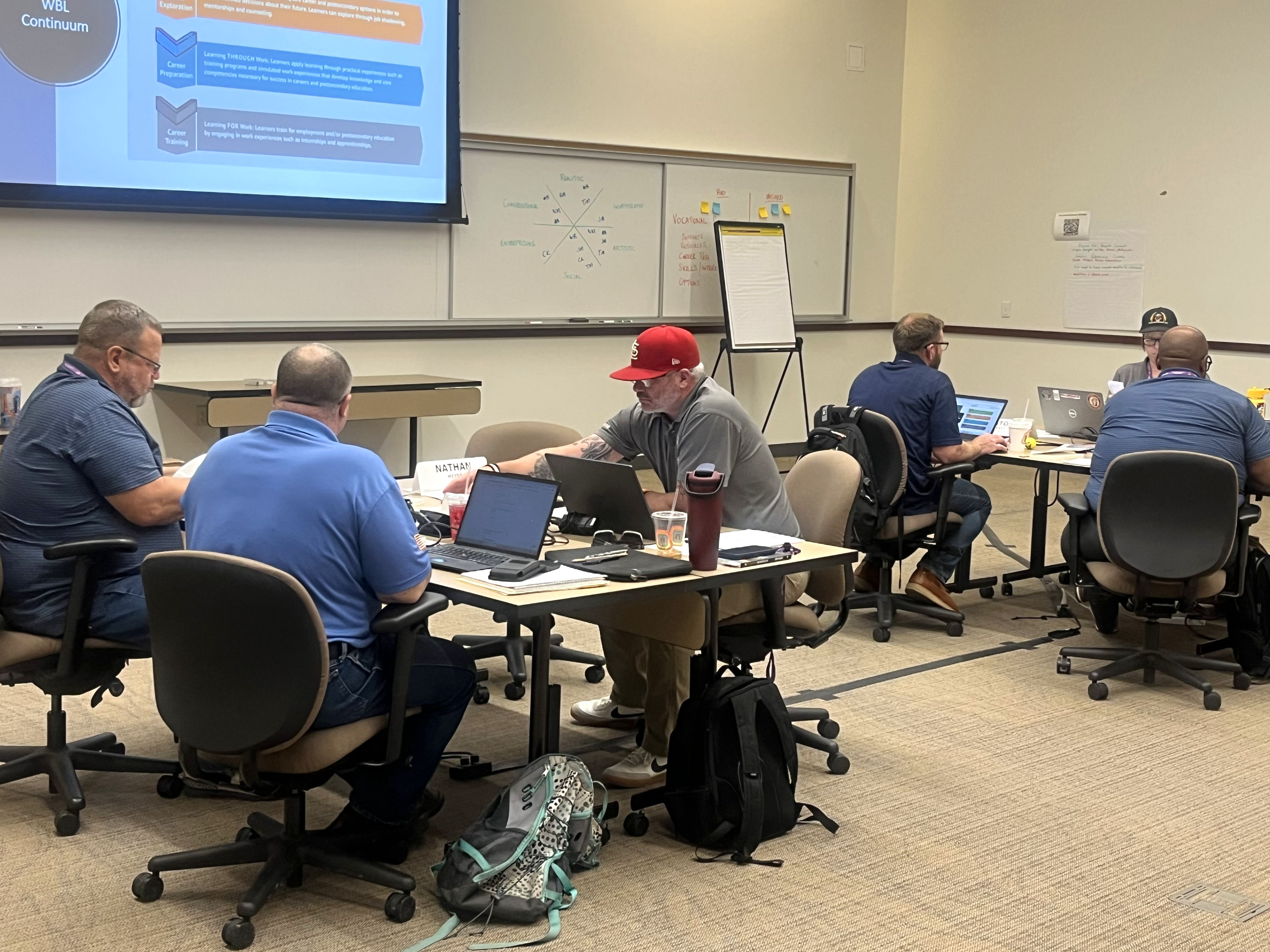 Two tables of people are seen in a large classroom. Three people are sitting at each table. At the front of the room, there is a whiteboard with writing on it and a projection screen. The people are working on laptops.