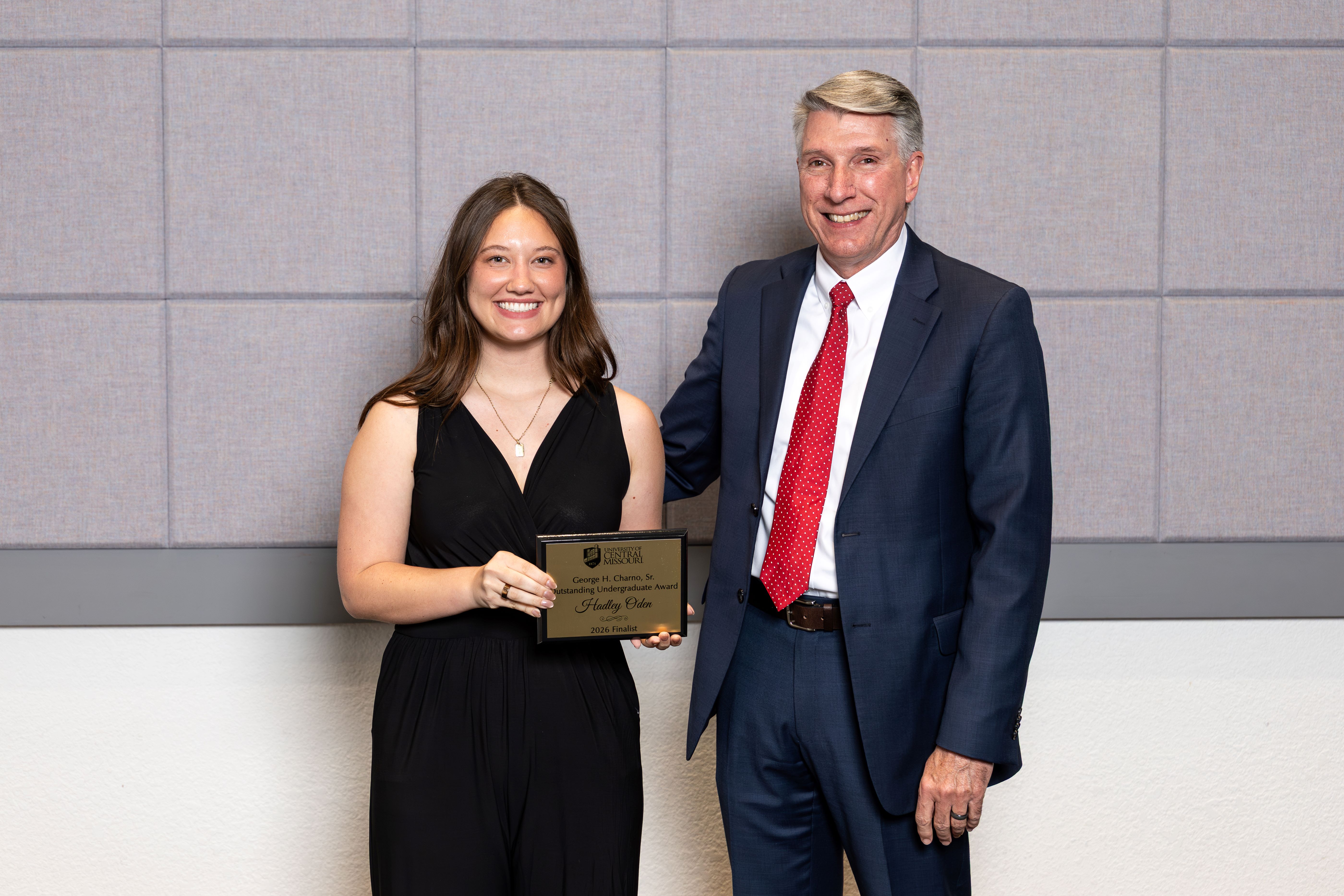 Charno Award recipient Hadley Oden stands next to UCM President Roger Best. Hadley is wearing a black outfit and holding a gold award plaque. Roger is wearing a navy suit and a red tie. They are standing in front of a gray wall.
