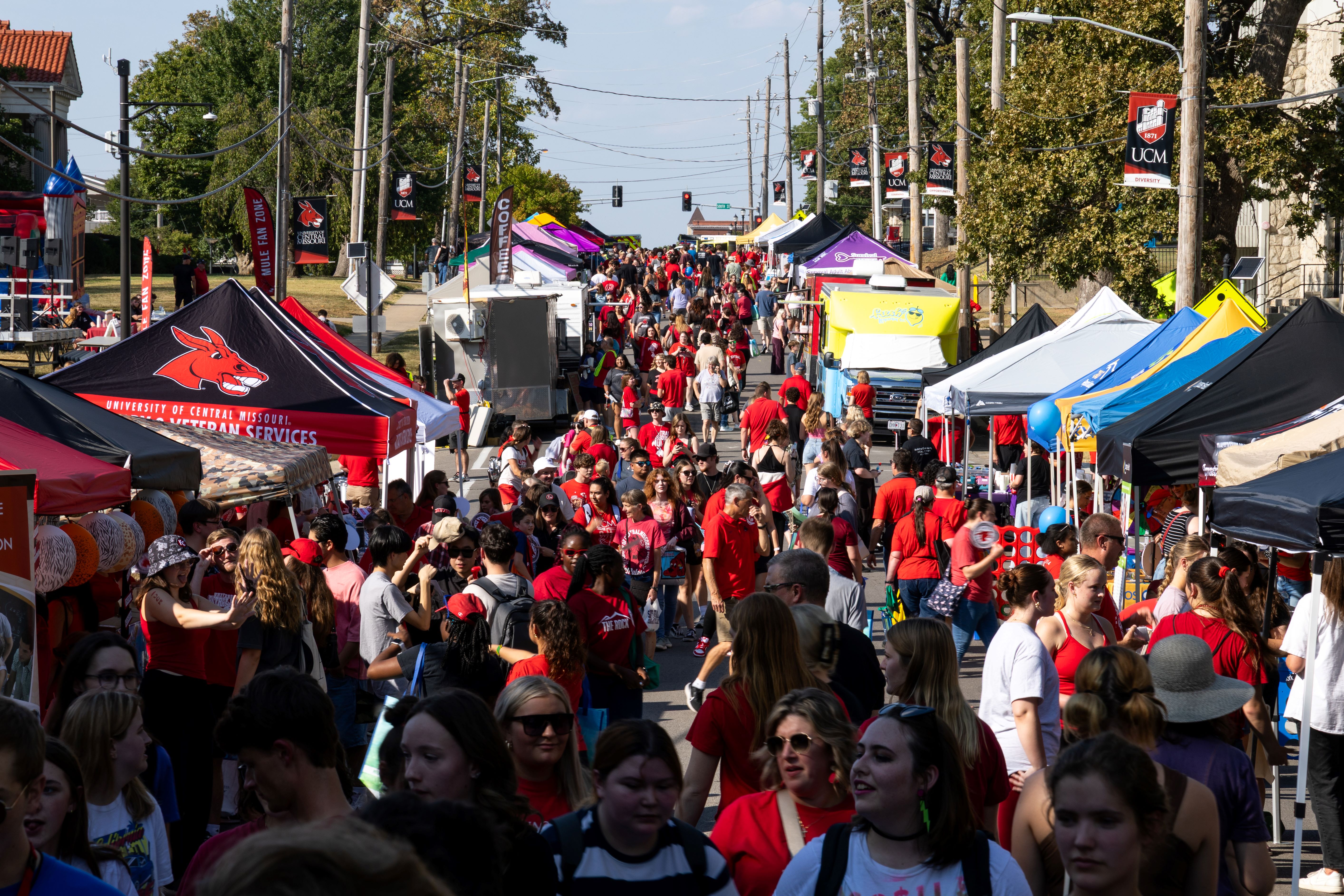 South Holden Street is lined on either side with dozens of pop-up tents in various colors. Hundreds of people are seen in the street talking, looking at vendors and walking. The crowd stretches all the way down the street. It is a sunny day.