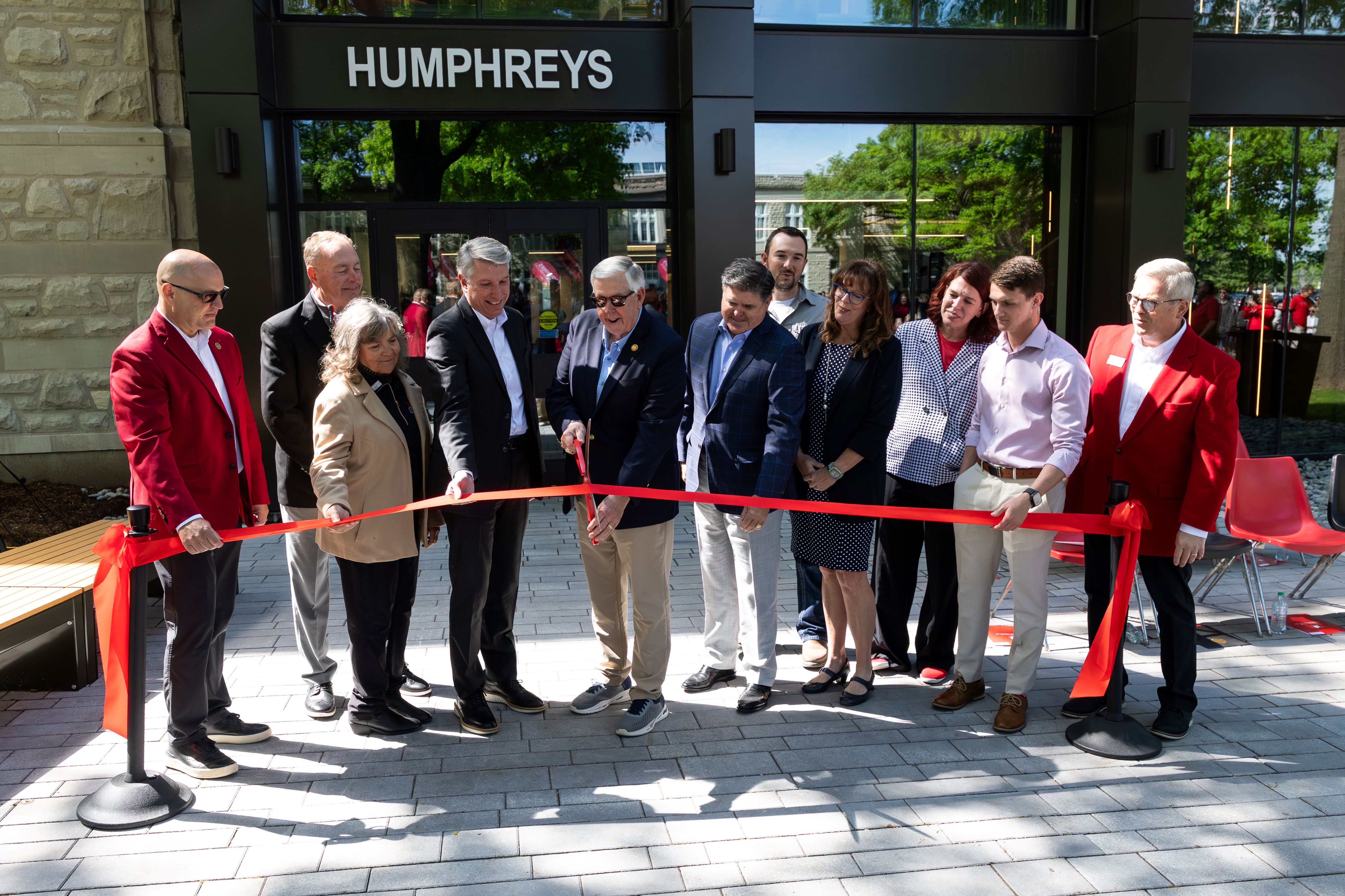 Eleven people stand in front of the Humphreys Building as the man in the center uses large scissors to cut a red ribbon.