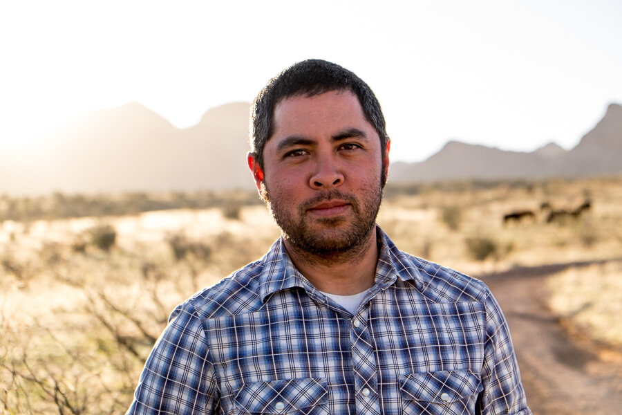A man is pictured from the shoulders up, with desert and mountains behind him