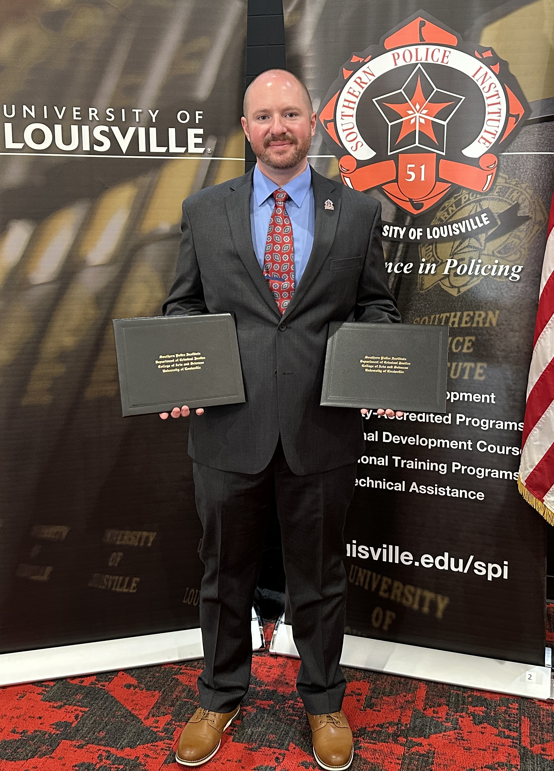 A person is wearing a dark-colored suit and holding two diploma/award covers. The person is looking at the camera and standing in front of two banners advertising the Southern Police Institute.