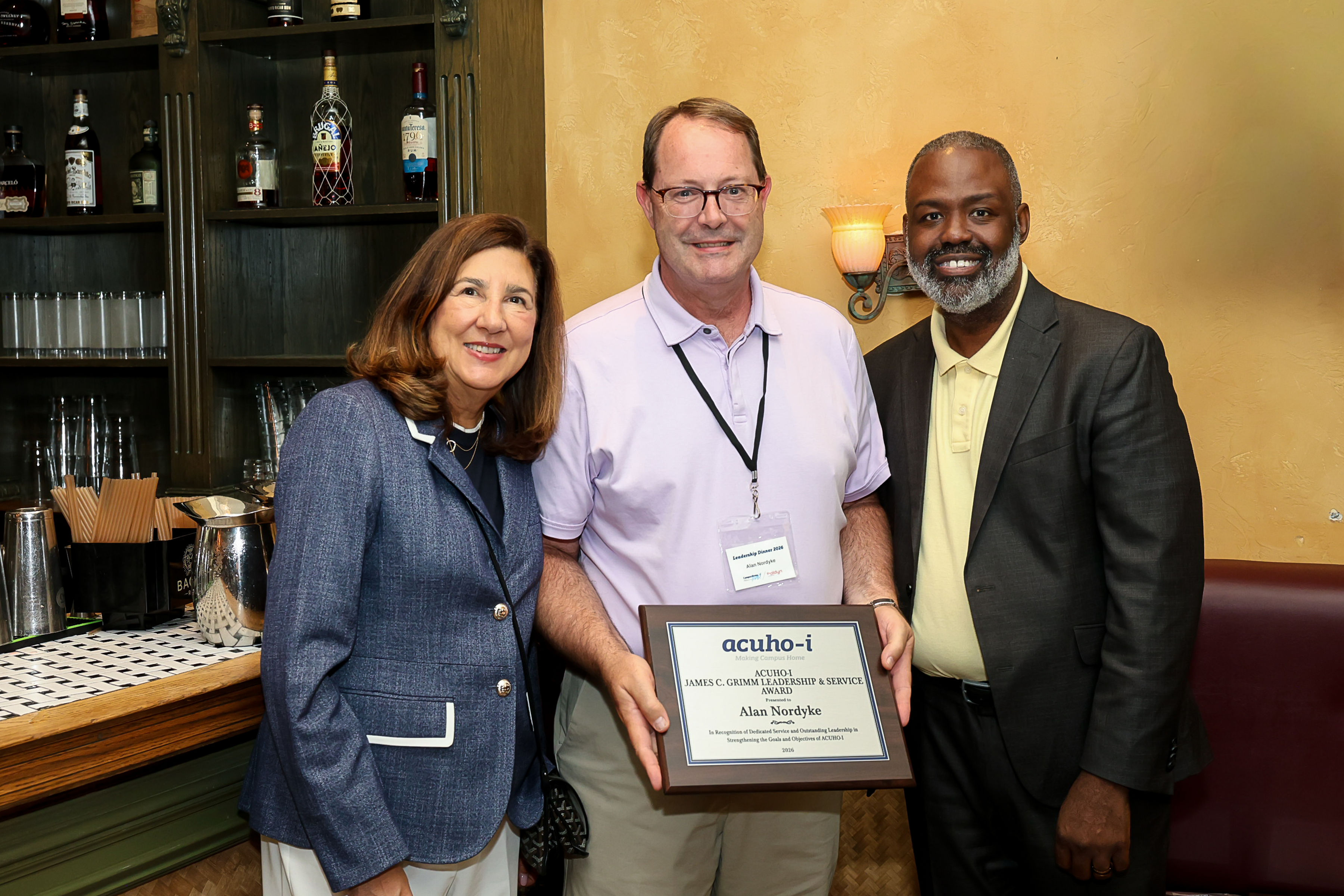 Three people stand next to each other in front of a bar in a restaurant. The award winner, Alan Nordyke, is in the middle wearing a lavender polo and khaki pants and holding an award plaque. The person on the left is wearing a blue blazer and white pants, and the person on the right is wearing a dark gray suit jacket and pale yellow polo.