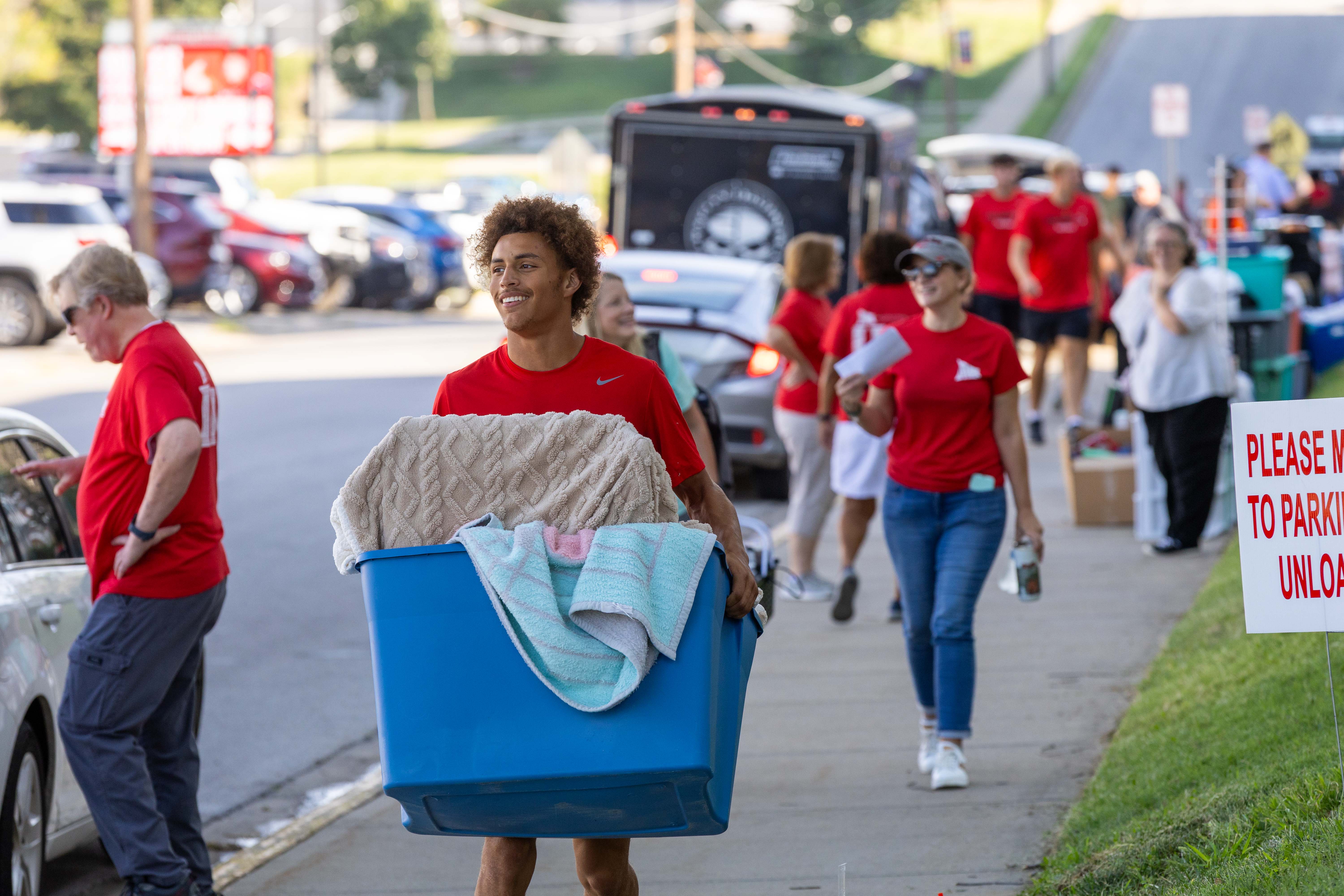 In the foreground, a person wearing a red UCM T-shirt is carrying a big blue plastic storage tote and towels can be seen draped over the side. The person is walking on a sidewalk. Behind the person, several parked cars and many people are seeing unloading items from the vehicles.