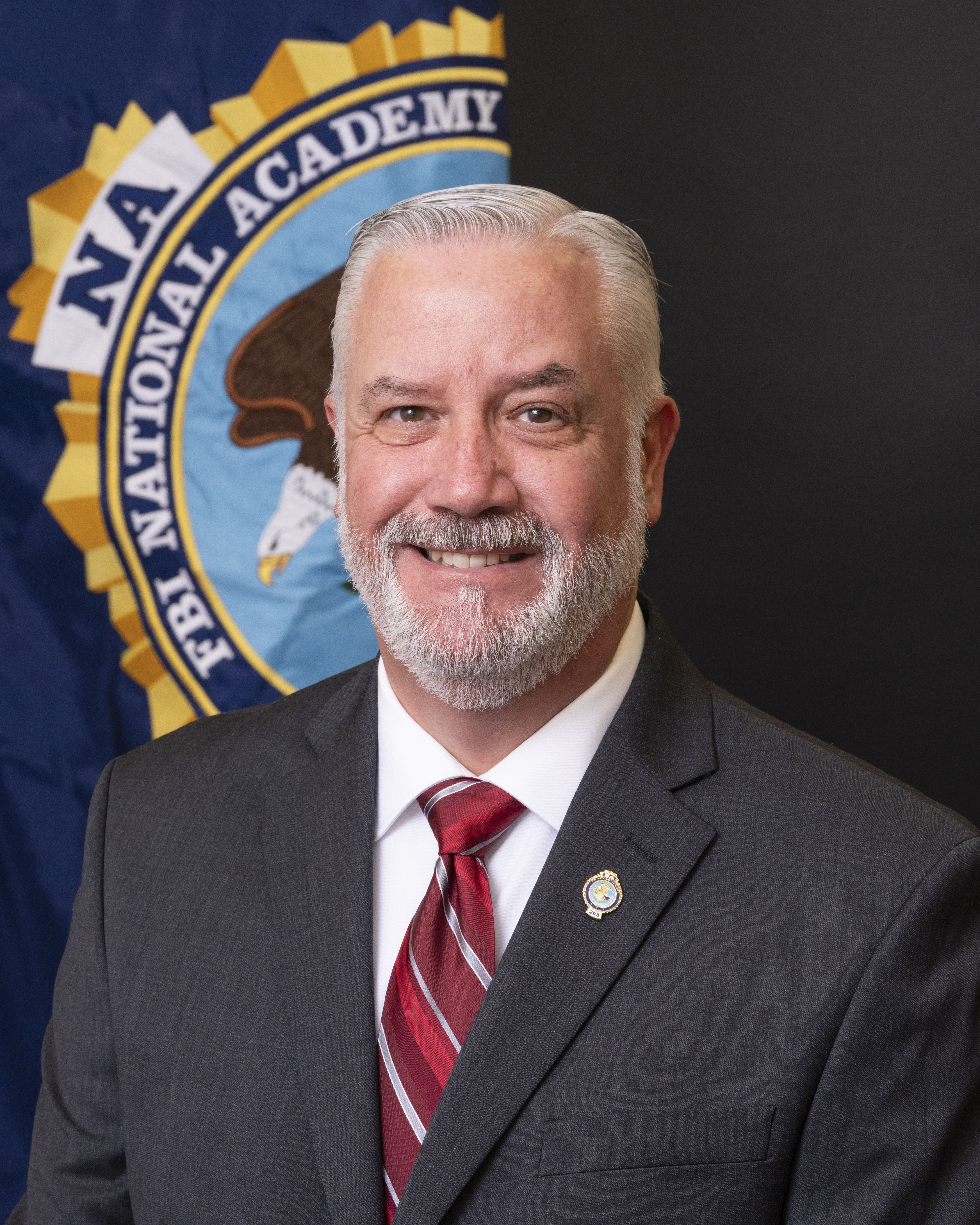 A person is seen in a professional portrait from the shoulders up. He is wearing a dark gray suit with a white shirt and red striped tie. Behind him, the FBI National Academy seal is partially seen.
