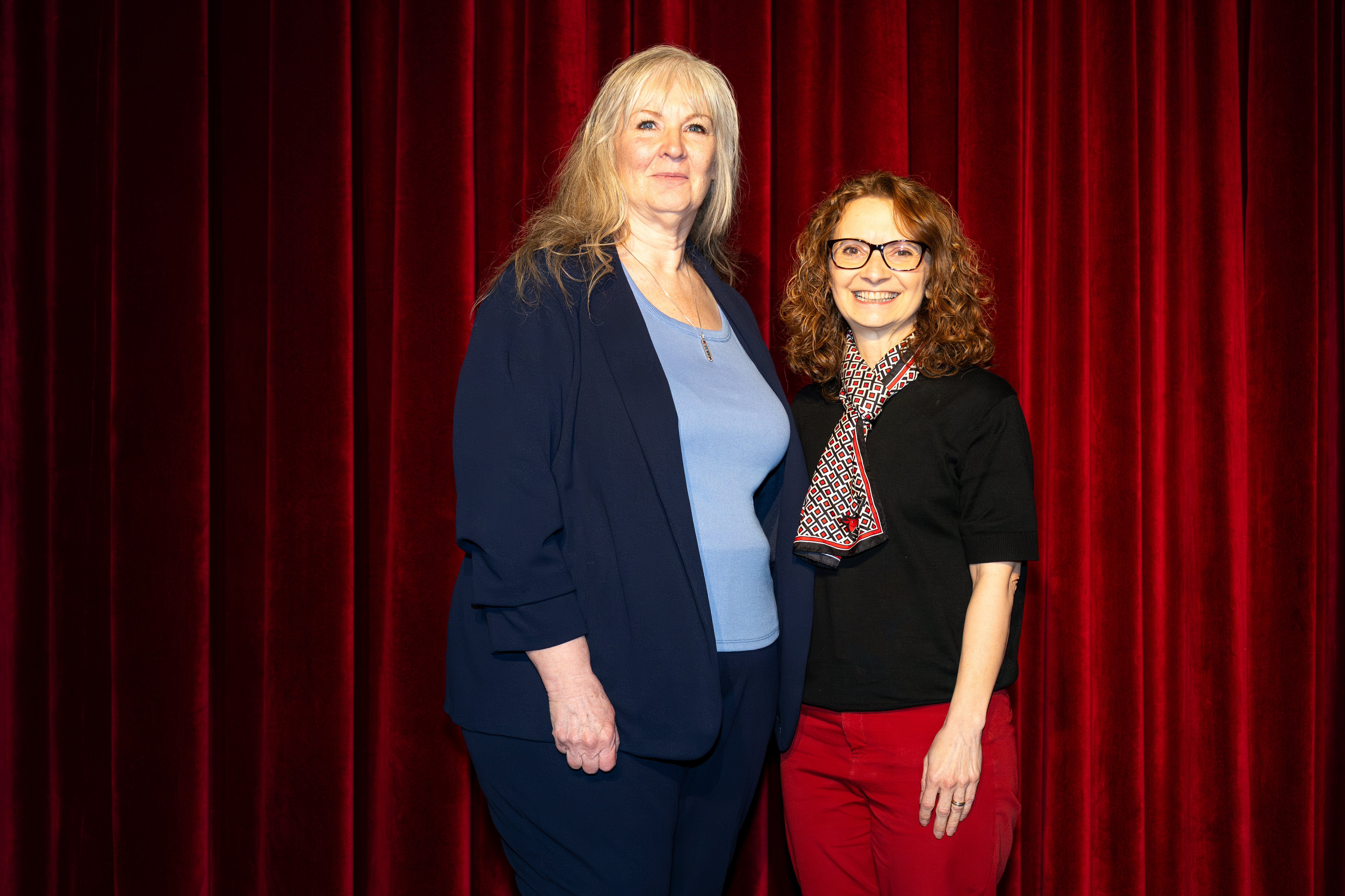 Two people stand next to each other in front of a red curtain. They are looking at the camera and posing for the photo.