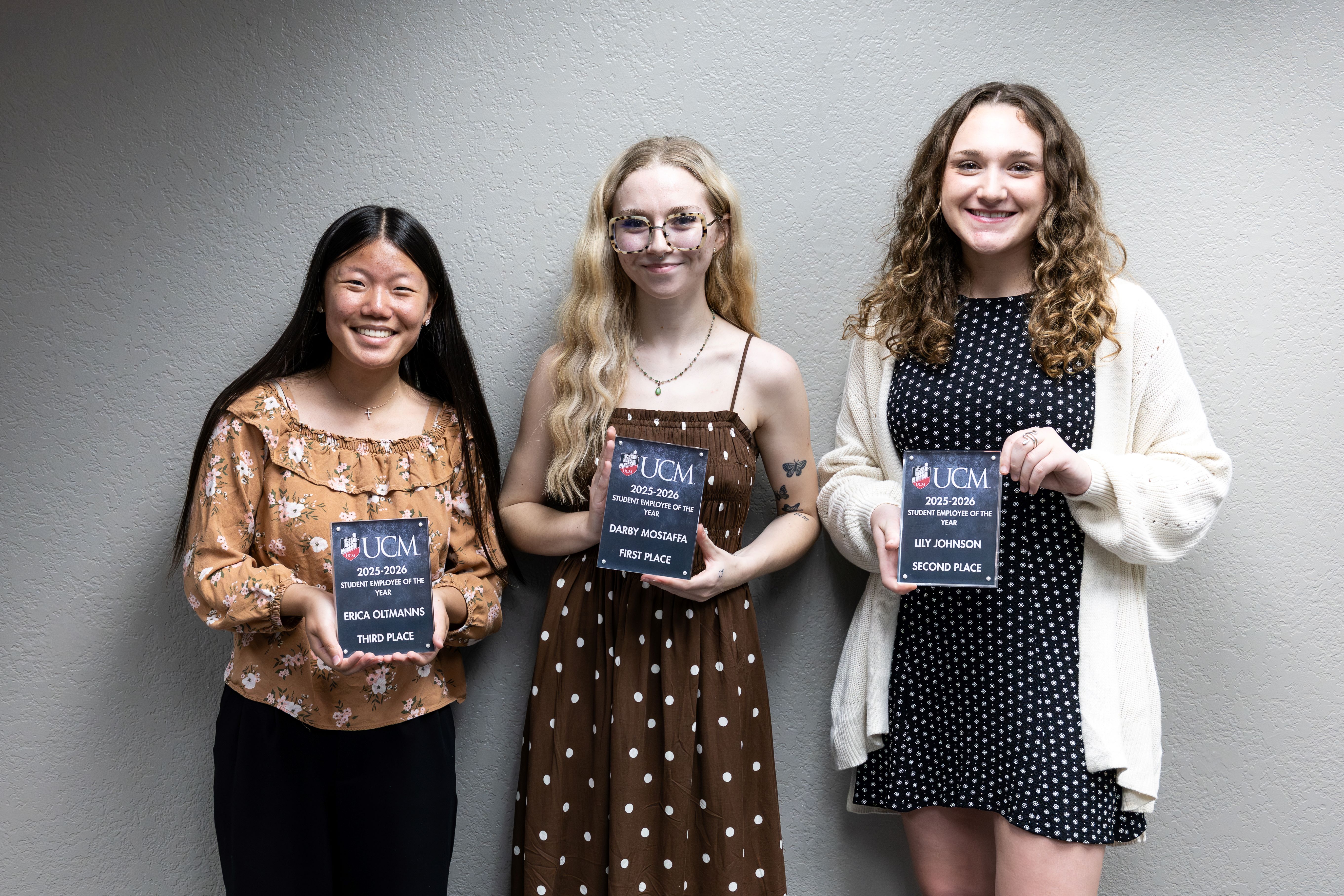Three students stand side by side in front of a gray wall. Each is smiling and holding a Student Employee of the Year award plaque.