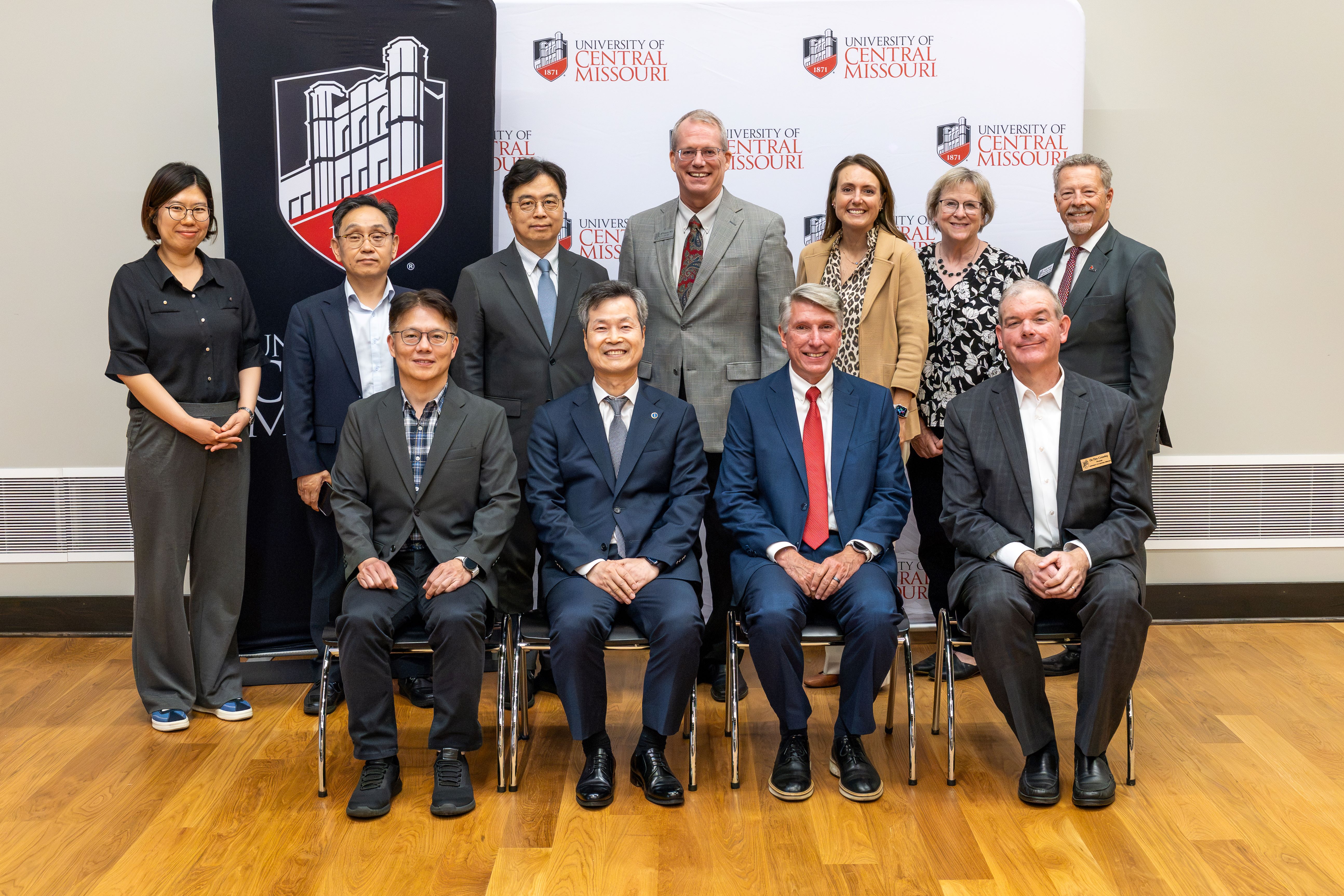 Four people wearing suits are sitting next to each other in black plastic/silver metal chairs. Behind them, seven people stand in a line, all wearing suits and business attire. The entire group is standing in front of a white backdrop with the UCM logo and 